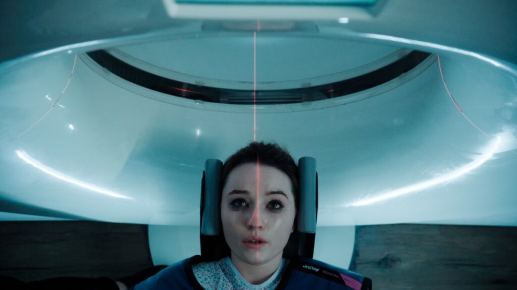 Belle Gibson (grana przez Kaitlyn Dever) leży w urządzeniu medycznym, a laserowa wiązka skanuje jej twarz. Scena ma futurystyczny, chłodny nastrój, podkreślający atmosferę tajemnicy i niepokoju.