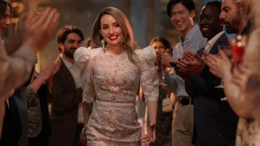 Belle Gibson (Kaitlyn Dever) uśmiechnięta, ubrana w elegancką białą sukienkę z koronką, przechodzi przez tłum ludzi, którzy biją jej brawo. Scena przedstawia moment jej triumfu i medialnego sukcesu.