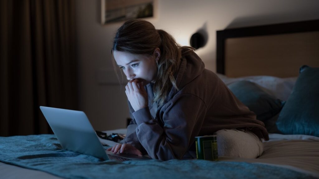 Belle Gibson (Kaitlyn Dever) siedzi na łóżku w przyciemnionym pokoju, wpatrując się w ekran laptopa z wyrazem skupienia i zmartwienia. Obok znajduje się otwarte opakowanie tabletek, co sugeruje moment refleksji lub wątpliwości.